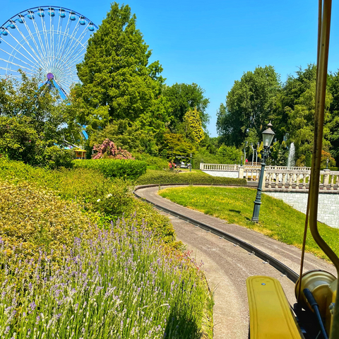 View from a car of Les Tour des Jardins car ride at Walibi Holland. Ferris wheel at top right. Green trees, bushes and lavender. Lamp post, bridge and fountain visible on the right. 