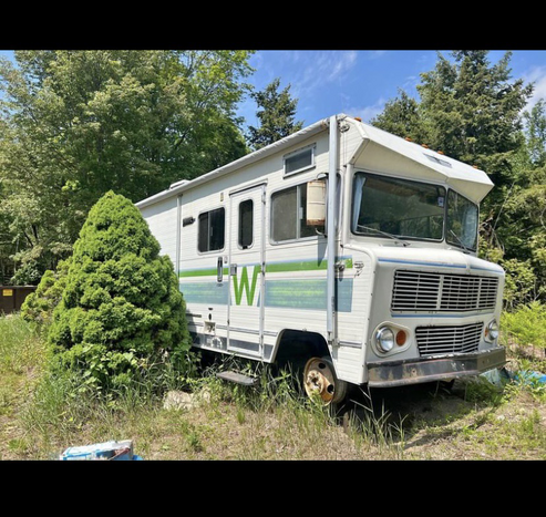 Old Winnebago RV sitting in a wooded area on a sunny day.