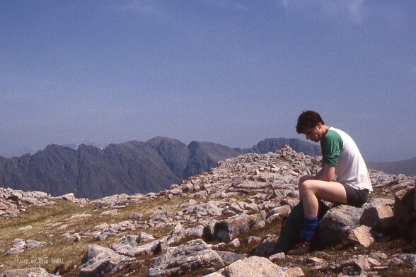 Young man sat on a rocky hill summit, wearing t shirt and shorts. A rocky ridge is visible in the background. Sunny with blue sky.