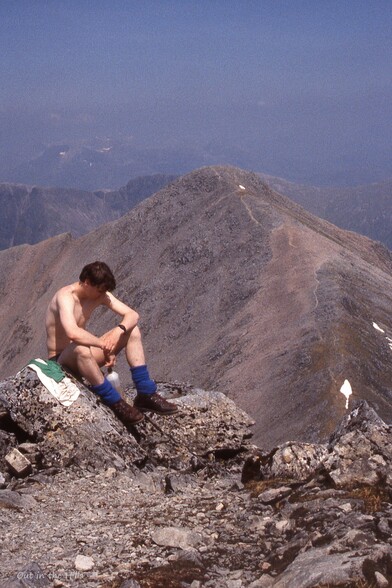 Young man, now topless, sat on a rocky ridge, which drops then ascents to a summit at the back. Blue sky and sunny but hazy