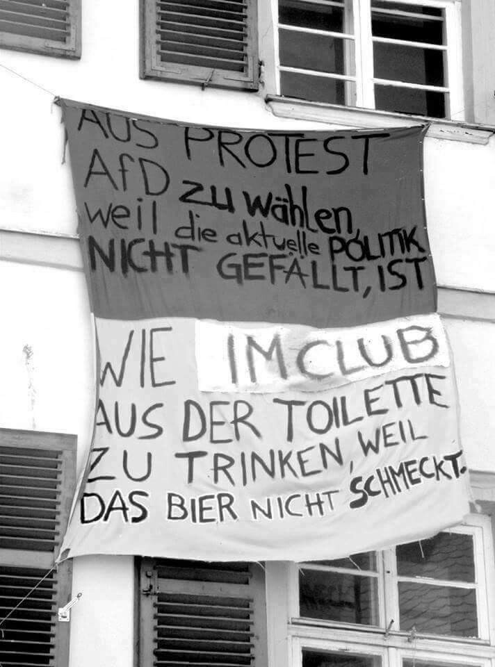 !B Schwarz-weiß Bild Banner an einer Hausfront mit der Aufschrift "Aus Protest AfD zu wählen, weil die aktuelle Politik nicht gefällt, ist wie im Club aus der Toilette zu trinken, weil das Bier nicht schmeckt!"