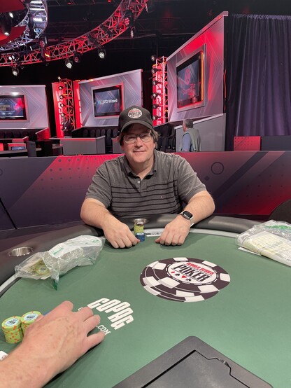 A man in his fifties sitting at a one of the finals tables at the World Series of Poker with a small stack of chips in front of him. He wears a cap and a gray shirt. 