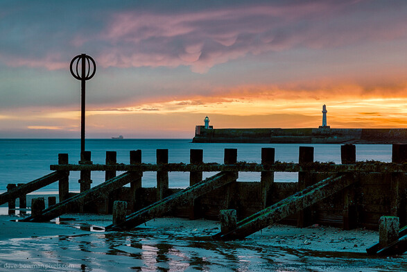 In this photograph capturing the tranquil essence of Aberdeen harbour at sunrise, your imagination can transport you to a peaceful coastal haven. The breakwater stands tall in the distance, its presence offering a sense of security and stability. In the foreground, a beach groyne and marker create elegant silhouettes against the awakening sky. A ship gracefully glides across the vast expanse of the sea, a mere dot in the distance, emphasising the grandeur of the ocean. The sky boasts a warm orange glow, casting a soft light that harmonises with the serene atmosphere. Shades of blue and orange dominate the scene, creating a visually captivating contrast. As you explore this image with your mind's eye, immerse yourself in the tranquility and beauty of this serene moment.