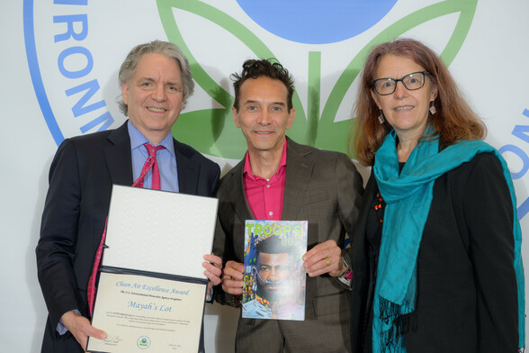 photo of three people in front of EPA logo. One white woman wearing a blue scarf and black dress, one latino man in a pink shirt and brown jacket holding the comic book Troops Run and one older white man holding the award certificate