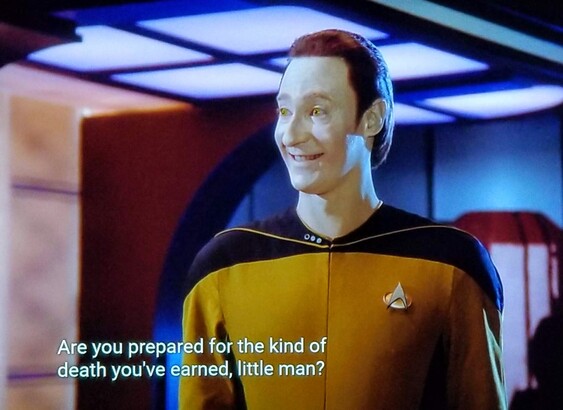 TNG scene. We're on ship, possibly in engineering?and looking at Commander Data. He's standing in golden uniform and has a bright and exaggerated smile on. Forehead is very tall, eyes beaming with intense cheer, kind of a horror movie type smile. Closed caption reads, "Are You prepared for the kind of death you've earned, little man?"