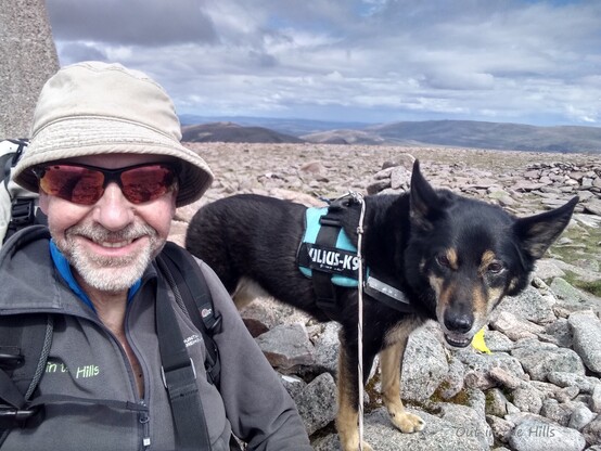Myself and Moray the dog looking at the camera, sat by a concrete pillar on a bouldery hill top.More rounded hills in the hazy distance.