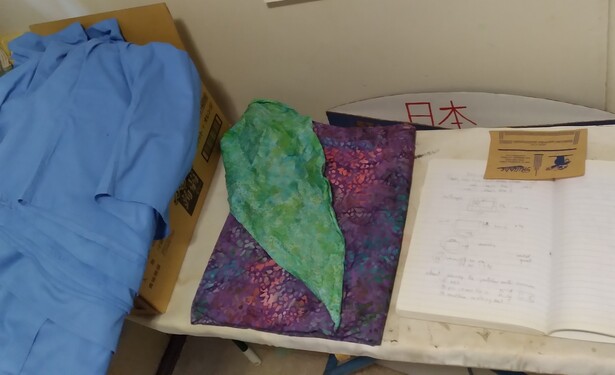 Photo of projects on an ironing board. Left to right: a pile of blue button down shirts; folded purple batik fabric with a green batik kerchief on top; composition book with pencil notes. Bag project not shown because it doesn't exist at all.