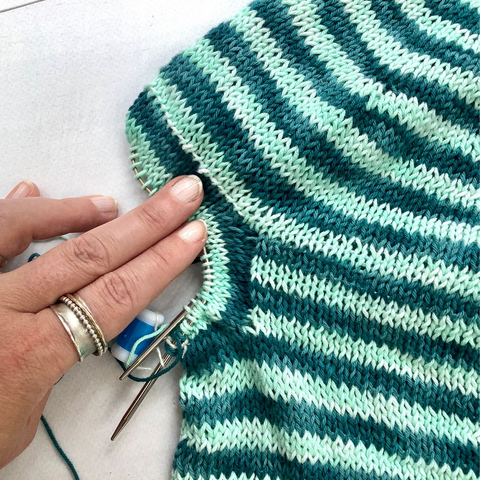 Flat lay view of the yoke and armhole of the back of a striped circular yoke garment in teal and light turquoise. The armhole stitches are placed on a circular needle. It is being held down by a person wearing a large silver fidget ring.