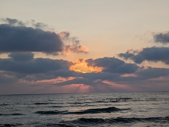 Sunset from the beach, the sun just behind the clouds last rays emitting from around the cloud in pink and purple. The cloud is gray and the light from above is orange. The water is almost empty except for a line of swimmers 