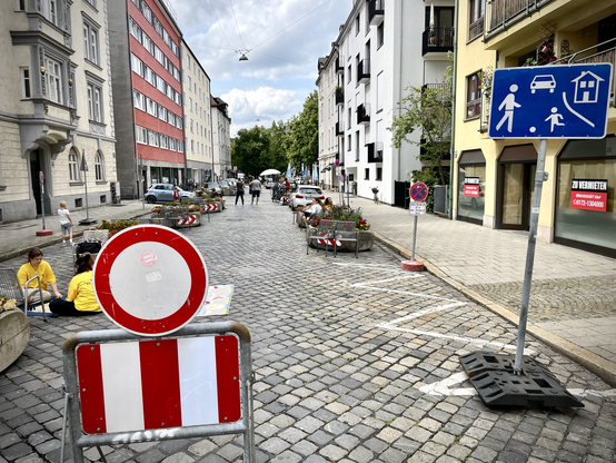 Für Eröffnung verlehrsberuhigter Straßenabschnitt mit Pflanzkübeln, Stühlen und Menschen. 