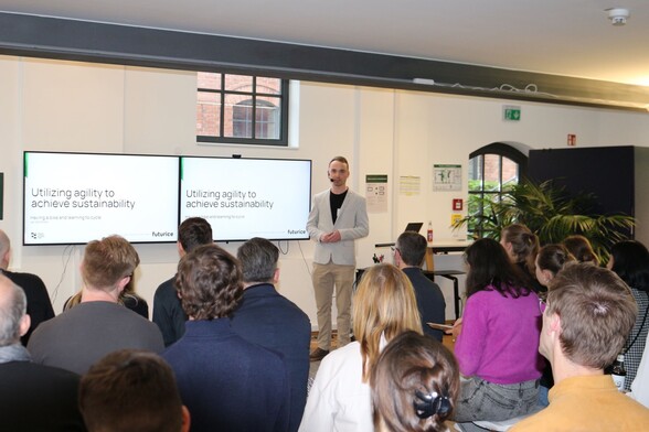 A person speaking in front of people sitting. Slides in the background saying: “Utilizing agility to achieve sustainability”