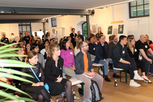 Many people sitting and standing in a room, looking towards the right and listening to a talk.