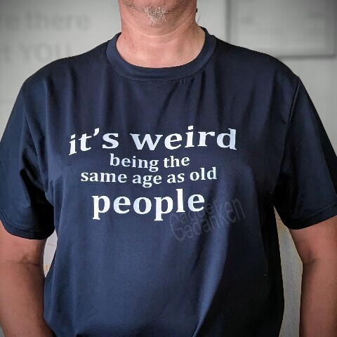 Person trägt ein dunkelblaues T-Shirt mit dem weißen Aufdruck: "It's weird being there same age as old people."