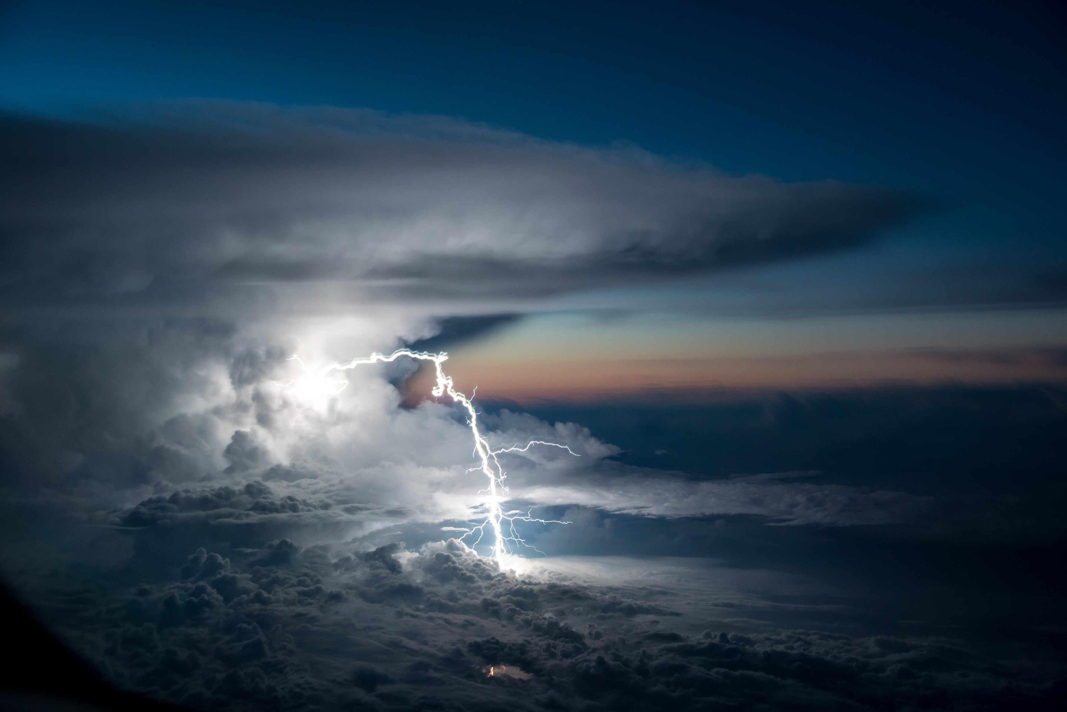 The view from a cockpit window shows a cumulonimbus incus cloud on the left of the frame, towering above the cloud ceiling. A writhing lightning bolt with many filaments arcs from the cloud's right, illuminating everything around it. The twilight sky <br />behind transitions from burnt oranges near the horizon to deep blues above