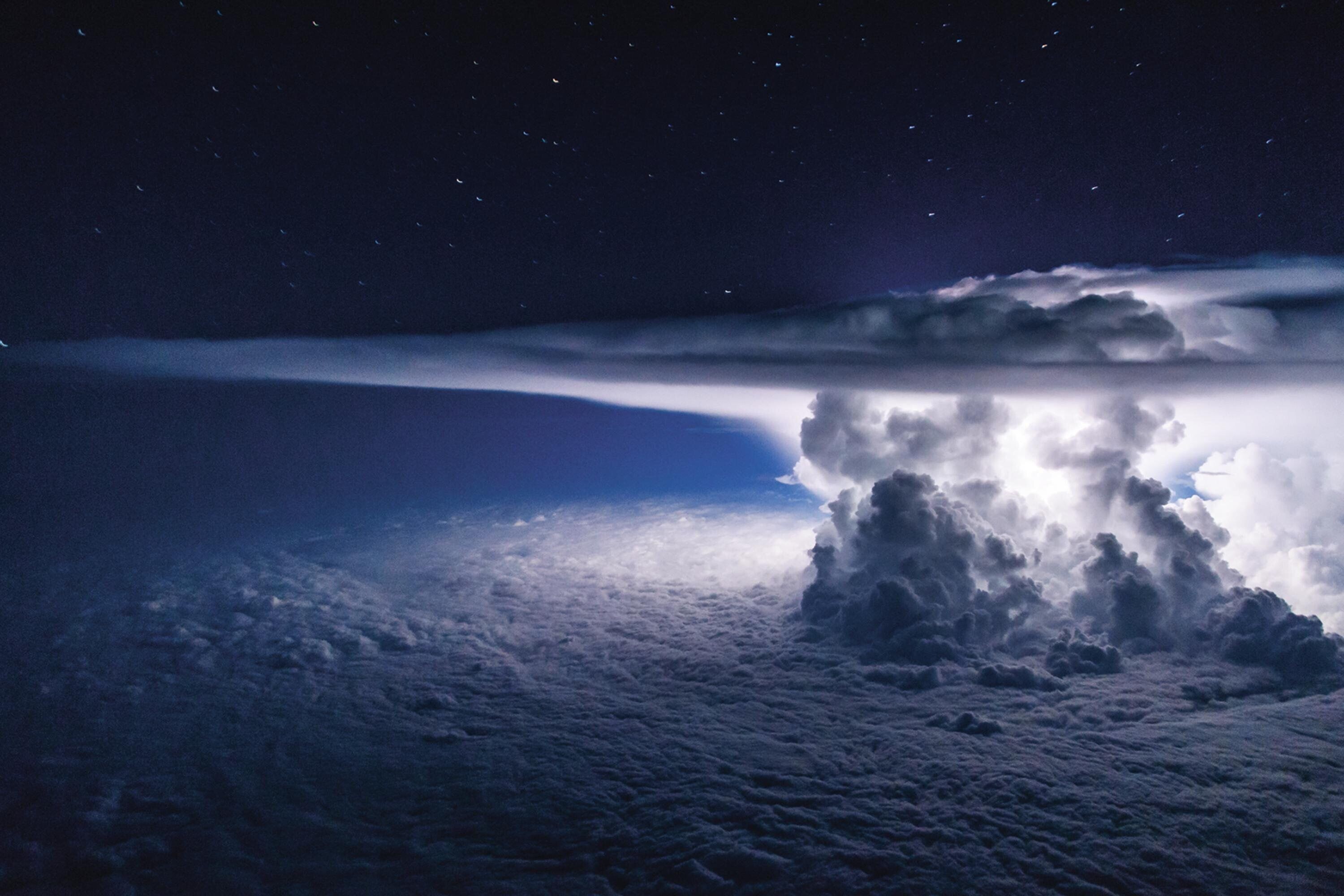 A towering cumulonimbus incus cloud appears on the right of the frame, illuminated from within by unseen lightning. It acts as a colossal lamp, filling the space between its "anvil" and the cloud ceiling with a ghostly glow. Above the cloud's "anvil," <br />stars sparkle in an inky evening sky.