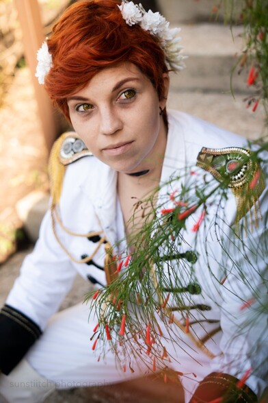 Portrait photo of me cosplaying Kiriona Gaia. I'm sitting, leaning forward and looking to the viewers right and there is a plant with tubular red flowers in the foreground.