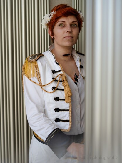 Portrait photo of me cosplaying Kiriona Gaia. In the background are metal bars pressed together which I am leaning against hands are down and I am looking to the viewers left