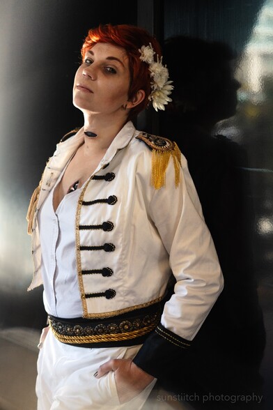 Portrait photo of me cosplaying Kiriona Gaia. Body is angled to the left and I have my hands in my pockets The lighting is very dramatic coming in from the left and back which is the direction I am turned I'm looking at the camera head tipped up. The background is dark and black