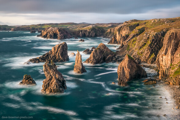In this captivating photograph, you can feel the serenity of the Mangersta Sea Stacks on the Isle of Lewis in the Outer Hebrides, Scotland, UK. The soft, grey sky serves as a tranquil backdrop, while the warmth of the setting sun gently illuminates the majestic sea stacks, casting a golden hue upon their rugged forms. The seawater below possesses a subtle, graceful motion, as if nature is slowly embracing the evening. With each touch of the breeze, you can almost sense the soothing sound of the waves dancing against the ancient rocks. A serene and breathtaking moment frozen in time, where you can almost taste the salt in the air and feel the embrace of nature's beauty.