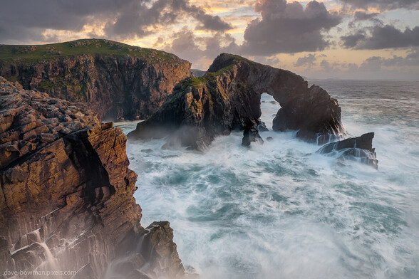 In this captivating photograph we encounter the majestic sea arch named Stac a' Phris on the Isle of Lewis in the Outer Hebrides, Scotland, UK. The arch stands proudly, gazing upon the restless North Atlantic, while the setting sun casts a golden glow that bathes the extraordinary rock formation. With turbulent motion in the sea water captured via a long exposure, the image conveys the dynamic nature of the surrounding waves. It is an awe-inspiring sight that transports you to the rugged beauty of this remarkable coastal landscape.