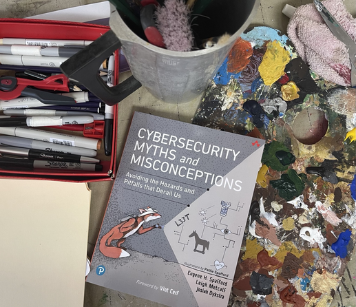 A copy of the book “Cybersecurity Myths and Misconceptions: Avoiding the Hazards and Pitfalls that Derail Us” on my studio workbench surrounded by pens, paper, a can of brushes, and a palette covered in dried paint.