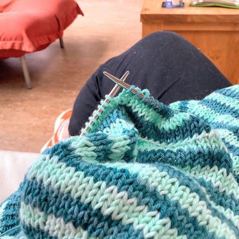 Closeup of some striped knitting on the lap of someone wearing black trousers and orange sneakers.