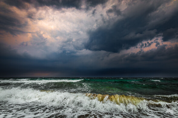 grau-blauer, wolkenverhangener Himmel, darunter grünes, anbrandendes  Wasser mit weißer Gischt.