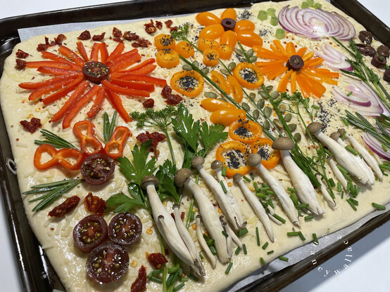Red and yellow bell pepper fireworks on focaccia bread dough, before baking. There are golden enoki mushrooms growing on the "ground" with snipped chives for grass. Bell pepper rings dusted with poppy seeds are the flowers. 