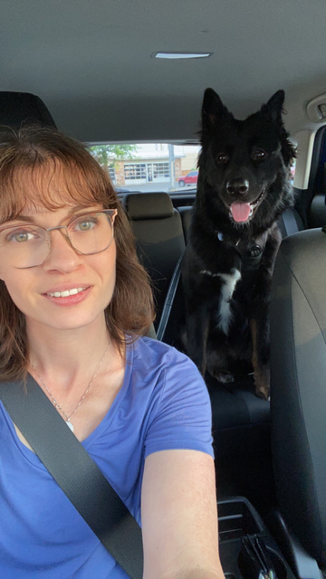 Woman with brown hair wearing a blue shirt sitting in a car with her black German Shepherd mix rescue dog in the back seat. 