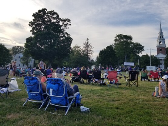 Concert-goers in lawn chairs on town common enjoying musical performance.