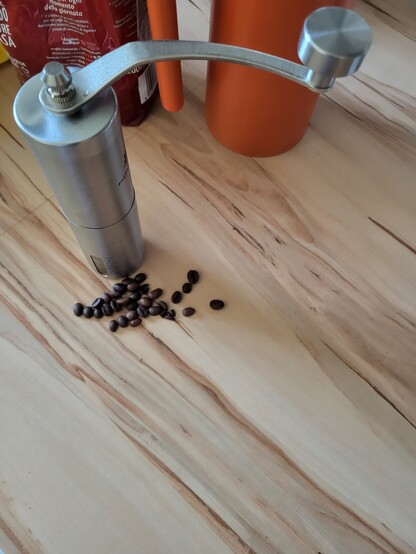 Eine schlanke metallene Dose mit Drehkurbel steht auf einem Holztisch. Davor liegen einzelne Kaffeebohnen. Im Bildhintergrund ist eine rote Kaffeebohnenpackung und eine orangefarbene Kanne im Anschnitt erkennbar.