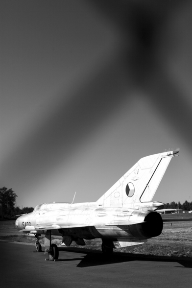 Parked fighter jet behind chain link fence. Black and white. Shot from the rear left of the plane.