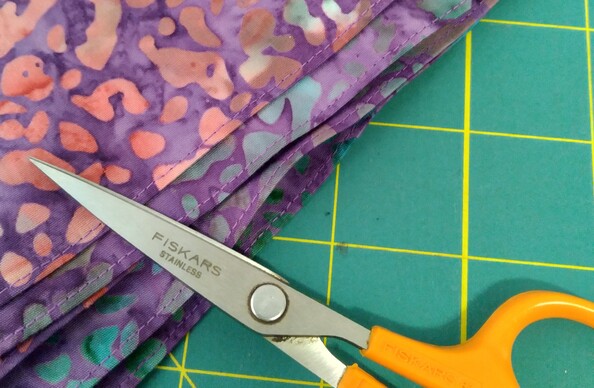 Close-up photo of bandana edges. Stack of purple batik bandanas on a green cutting mat, with a small pair of orange Fiskars scissors next to them.
