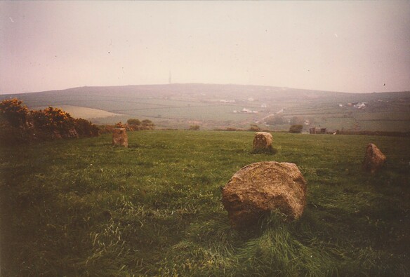 Colour photograph of the remaining stones of the south-east Nine Maidens Stone Circle. Four small stones lie along an arc in a green field on a slightly misty day. Behind is a hill with a few scattered farms and cottages and with a transmitting tower on the peak.