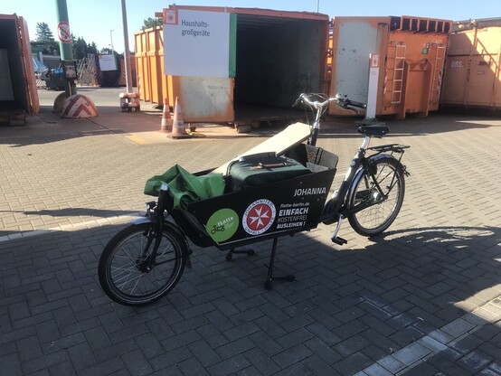 Ein Lastenrad bepackt mit Sperrmüll steht auf einem Parkplatz, im Hintergrund ein großer Recyclinghof-Container
