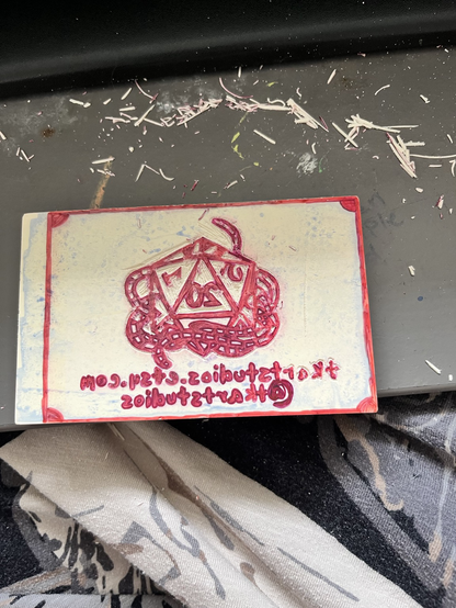 A hand carved stamp in progress. A reversed image of a d20 with yarn wrapped around it and the words TKArtStudios dot Etsy dot com and at TKArtStudios 
