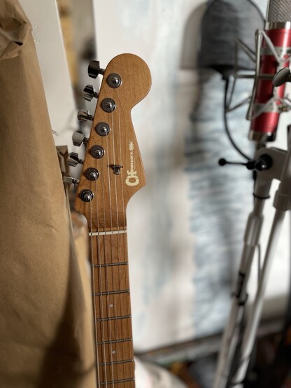 A close up of Nimona’s neck, my new Charvel DK 24 Pro Mod HH electric guitar. The neck is roasted/caramelized maple. It is full of wood character with swirls of darker lines throughout. The color is like a perfect warm tan, and the logo is in soft ivory. The tuners are Charvel labeled and silver. Substantial and nice. The nut is graph tusq, an equivalent to bone. It is off white. the neck is all one piece.