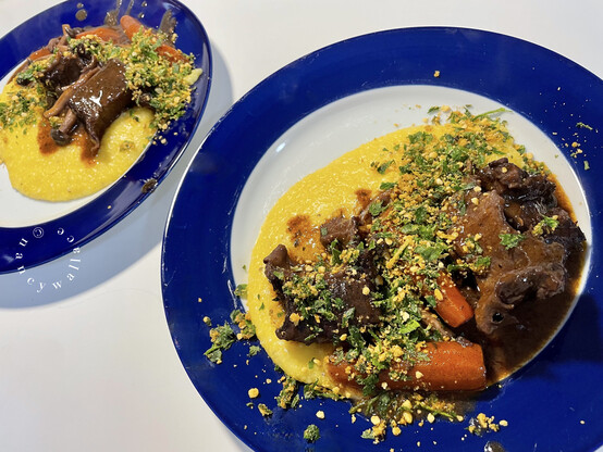 Two dinner plates: white with a blue rim. On each plate are some beef bourguignon short ribs with golden polenta for soaking up all the red wine juices. Carrots and shimeji mushrooms were also cooked with the dish. A parsley and lemon gremolata with toasted breadcrumbs is sprinkled over the top just before serving, as pictured. 