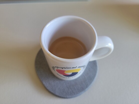 A white, 3 ounce, ceramic espresso (demitasse) cup with a shot of espresso, on a gray coaster on a white desk mat. The blue text on the cup reads "Cyber-Physical Systems Research Center" and wraps in a circle around the logo, which looks like a loosely interlaced, rounded triquetra with thick knots in solid red, yellow, and blue (each its own color).