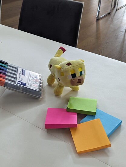 Auf den Tischen kann gemalt werden. Dafür sind sie mit Papier beklebt und ihr findet darauf bunte Klebezettel und Stifte. Und manchmal auch Kuscheltiere. Hier im Bild eine gelbe Katze aus Minecraft