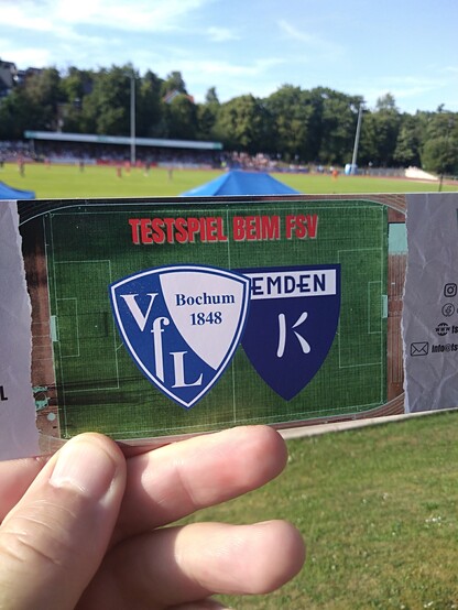 Empor gehaltene Eintrittskarte mir den Vereinsemblemen von VfL Bochum und Kickers Emden, im Hintergrund Sportanlagen und Waldrand.