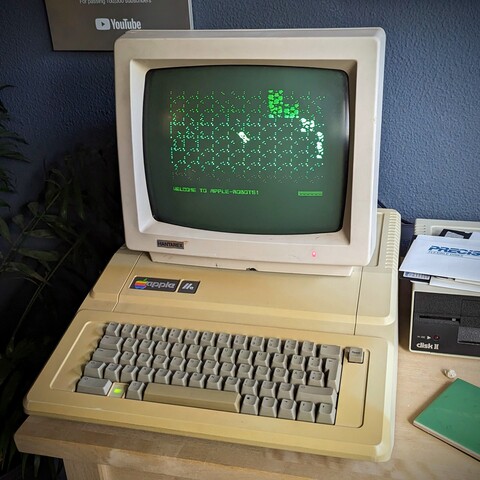 An Apple IIe, connected to a Drive II with some 5 1/4 disks on top of it, and the game Apple Robots on a vintage monitor.