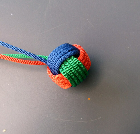 Pomme de touline : nœud à six faces en forme de boule.
Ici, chaque passage est effectué cinq fois.
La pomme est réalisée avec des fils de trois couleurs : rouge, vert et bleu.