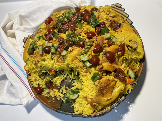 A serving dish showing a portion of the Upsidedown chicken has been removed. There are browned chicken pieces on the top, with golden rice, and a tomato-cucumber-parsley gremolata sprinkled over the top. Also: some toasted pine nuts for good measure.