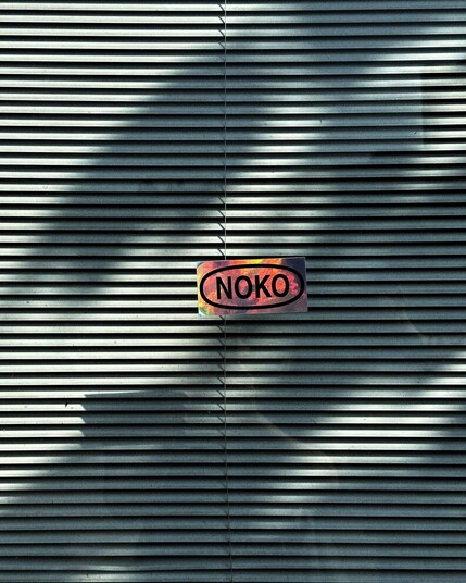 a rectangular chrome sticker with the text "NOKO" inside an outlined oval shape, hanging in front of what looks like a window with some closed blinds on the inside.