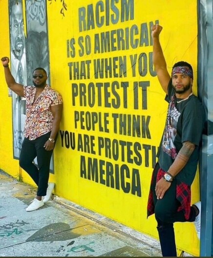 Two black activists are standing besides a poster, fist in the air.
The poster reads, in black letters on a yellow background: "Racism is so American that when you protest it, people think you are protesting America"

Alt description courtesy of @StrepsipZerg