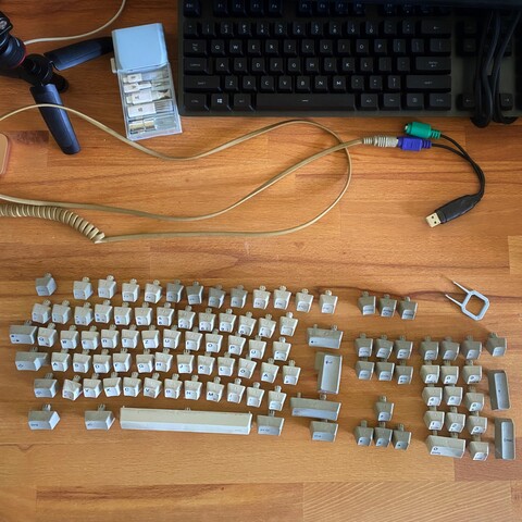 Detached IBM Model M keycaps in formation on a desk. Background: camera tripod, cleaning kit, another keyboard, coiled IBM cable with PS/2 connector and USB adapter.