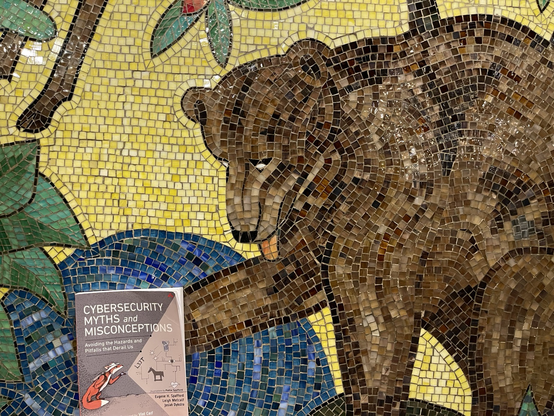 An image of a mosaic depicting a brown bear on a yellow background next to a pool of water. Next to the bear’s paw is a copy of the book Cybersecurity Myths and Misconceptions: Avoiding the Hazards and Pitfalls that Derail Us