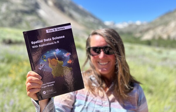 Allison holding the book "Spatial Data Science With Applications in R" from CRC Press by Edzer Pebesma and Roger Bivand in the foreground. On the cover of a book is an illustrated wildland wizard (a la Radagast) holding a staff while controlling a number of spatial data pieces (raster layers, polygons, projections) in the air. 