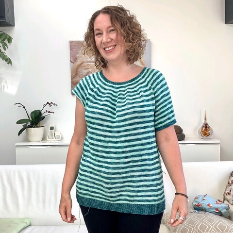 A woman with chin length curly hair smiles into the camera. She is wearing a Striped tee with only 1 sleeve.